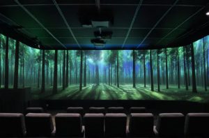 Image of a theater-like room with projections of a forest scene on 3 sides. There are comfortable chairs facing the front wall.